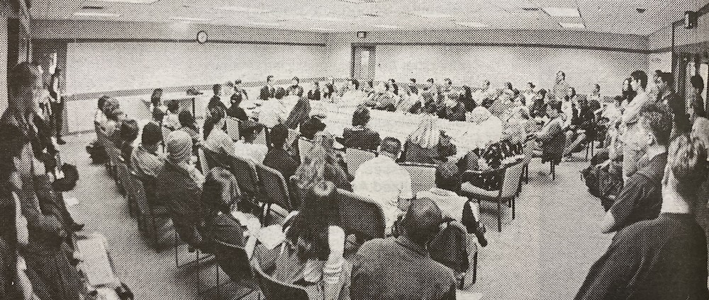 Show thumbnail preview A large group of people sits in a conference room with rows of chairs arranged in a semicircle around a central table.
