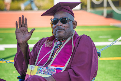 seated older man wearing graduation regalia and waving