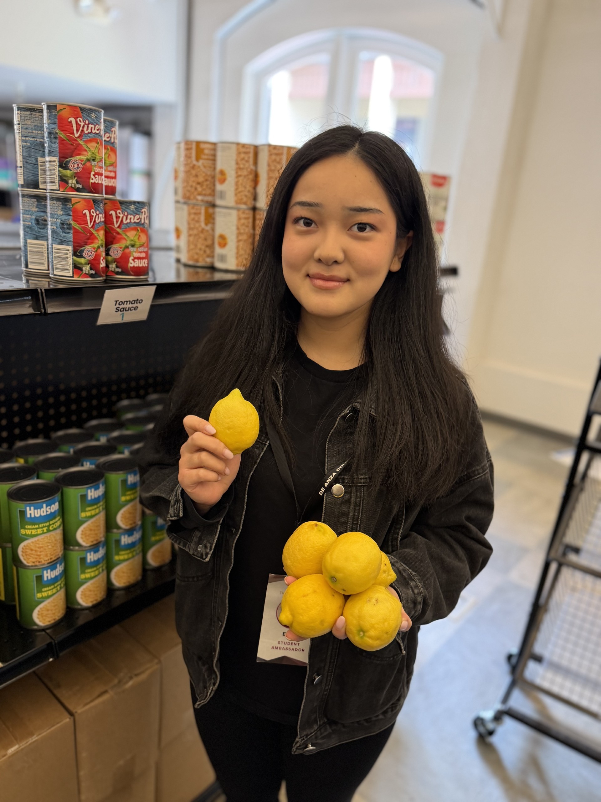 student wearing black is standing in front of shelves and is holding up lemons
